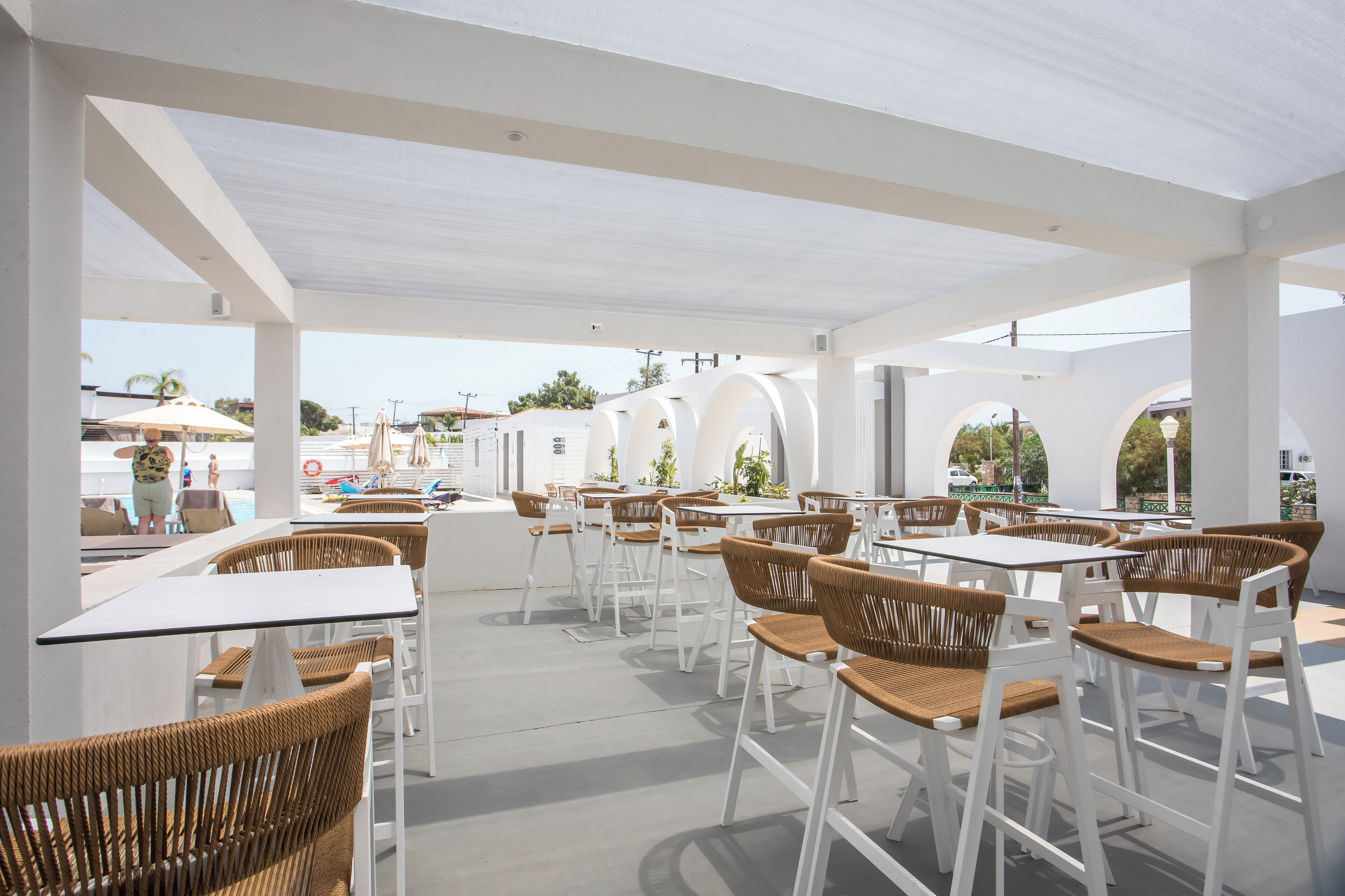 An outdoor cafe area with white tables and wicker chairs, featuring a clear view of the pool and palm trees in the background.