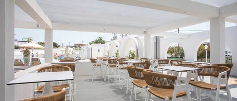 An outdoor cafe area with white tables and wicker chairs, featuring a clear view of the pool and palm trees in the background.