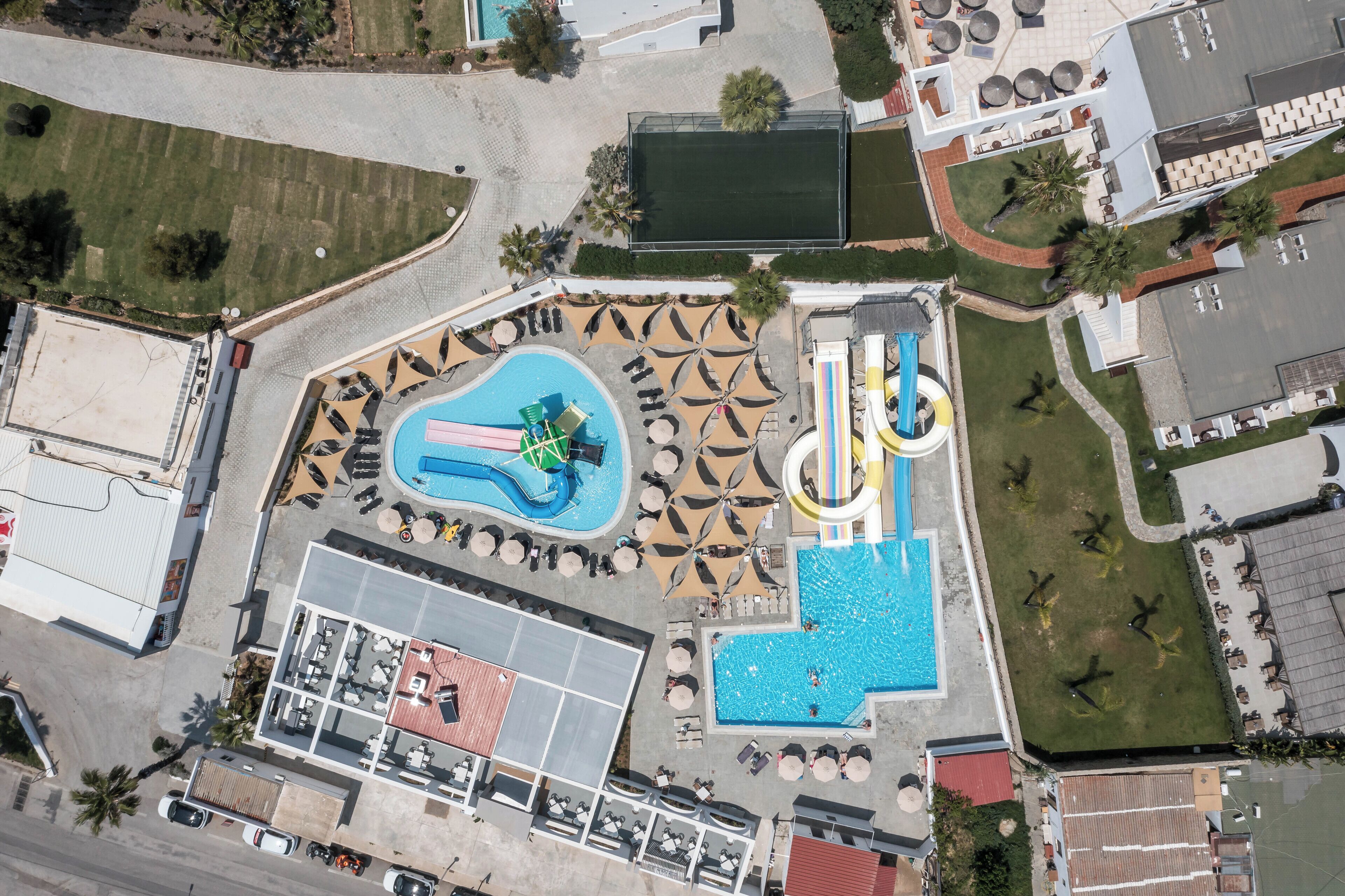 An aerial view of a resort with multiple swimming pools, a water slide, and a tennis court.