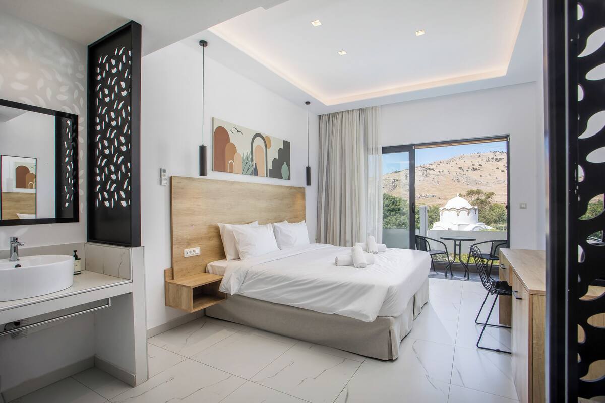 A modern hotel room with a large bed, a bathroom with a sink, a desk, and a balcony with a view of the hills.