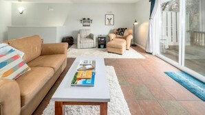 Ein Wohnzimmer mit einer Couch, einem Sessel und einem Couchtisch mit Büchern. Ein Schiebetürfenster führt in einen Außenbereich.