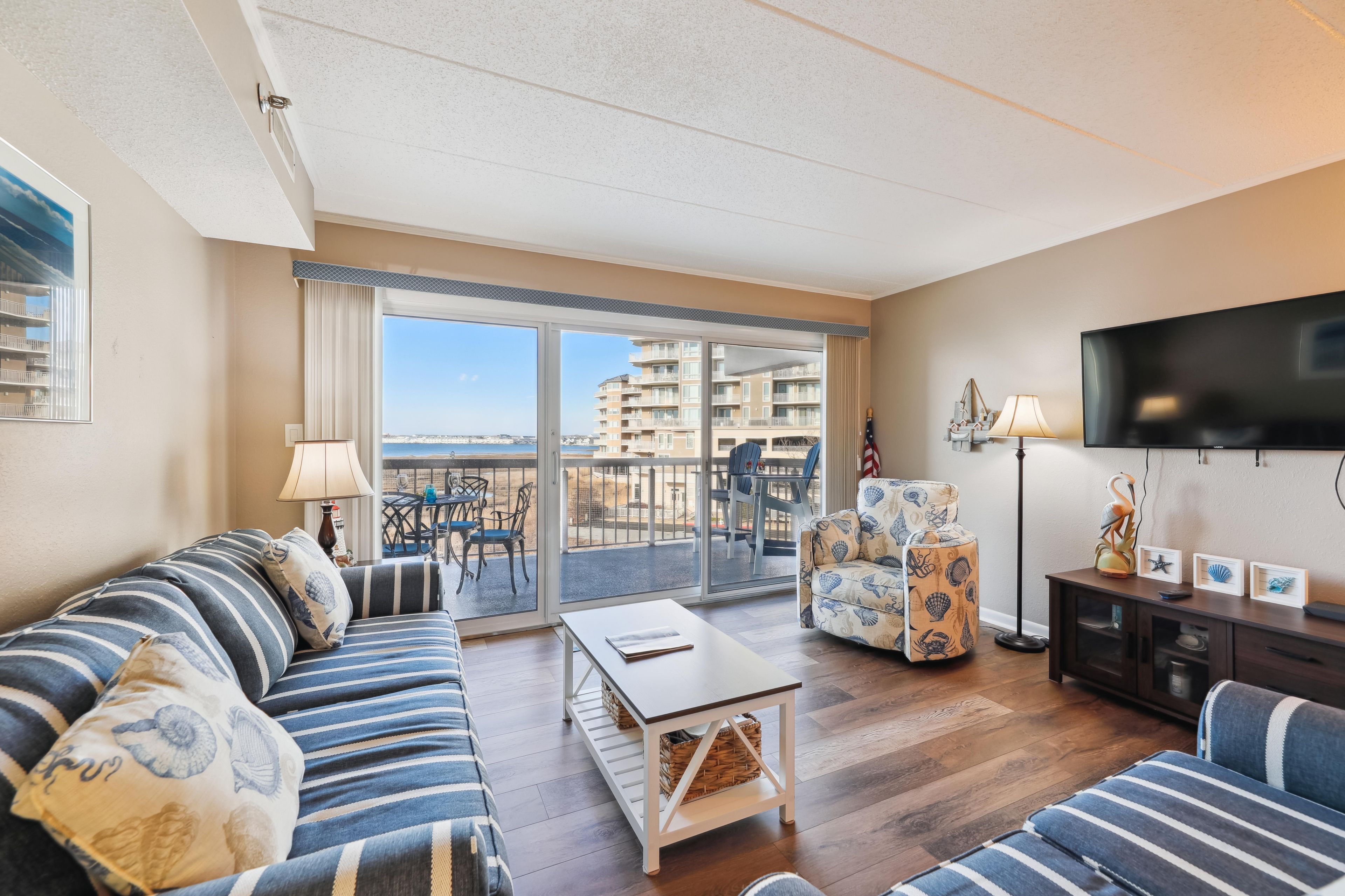 Een woonkamer met een bank, fauteuil en een tafel, een televisie en een balkon met uitzicht op gebouwen.