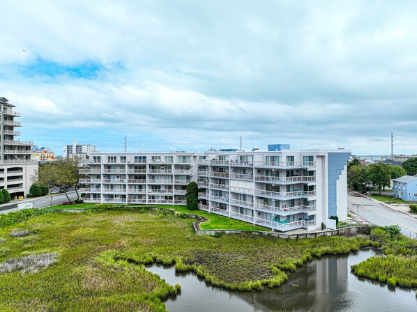 A modern multi-story residential building with balconies, a grassy area, and a small pond. - XPLORIE ACTIVITIES & LINENS INCLUDED*! Great 3BR bayfront condo on 79th Street with outdoor pool! (Ocean City)