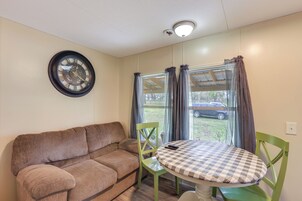 Quarto com sofá marrom, cadeira verde, mesa redonda com toalha xadrez, relógio de parede e janela com cortinas.