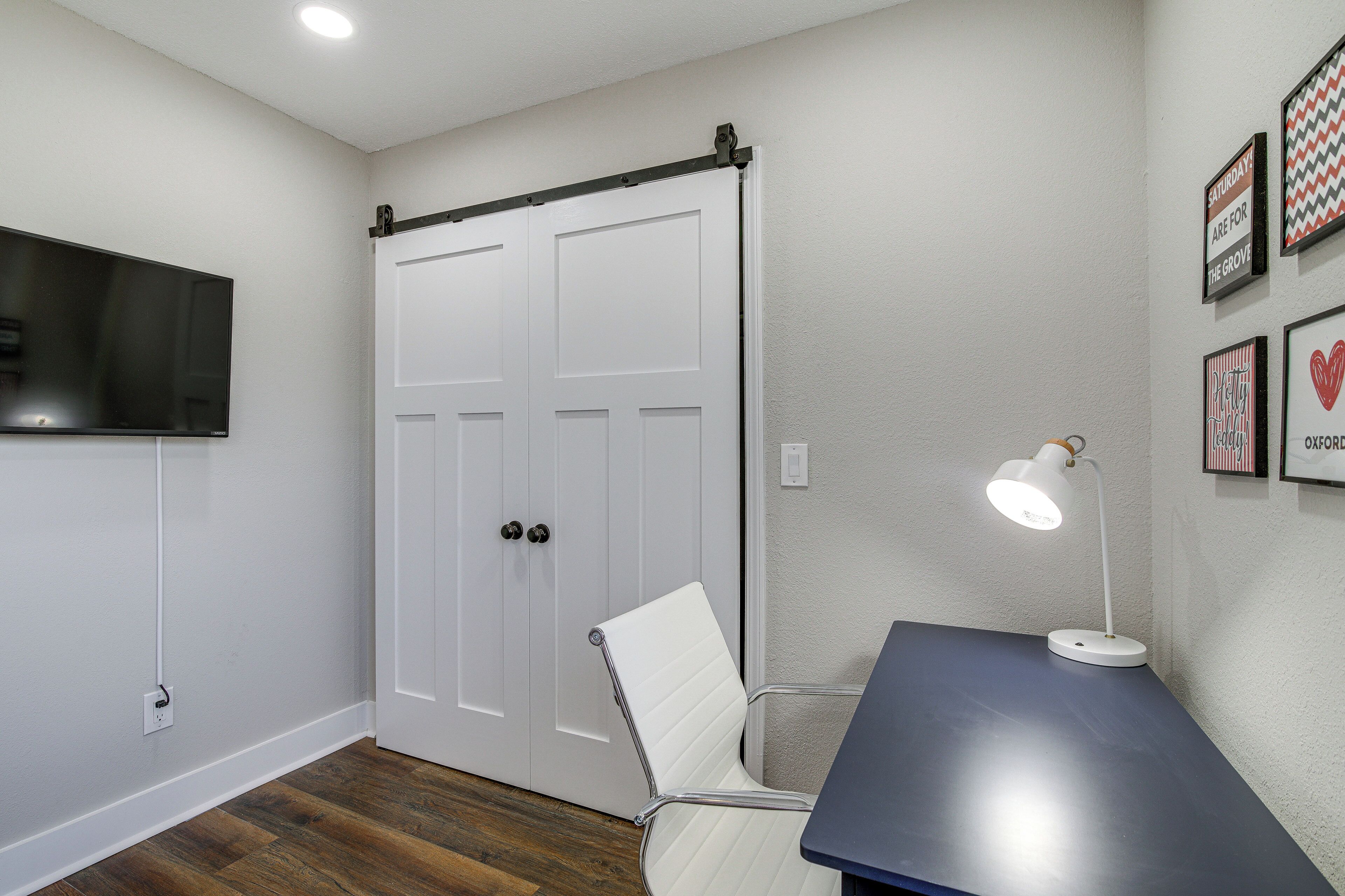 A room with a sliding door, a flat-screen TV mounted on the wall, a desk with a lamp, and framed pictures on the wall.