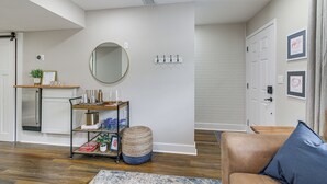 A living room with a brown leather sofa, a round mirror, a small round ottoman, a wooden side table, and a metal shelf unit with various items.
