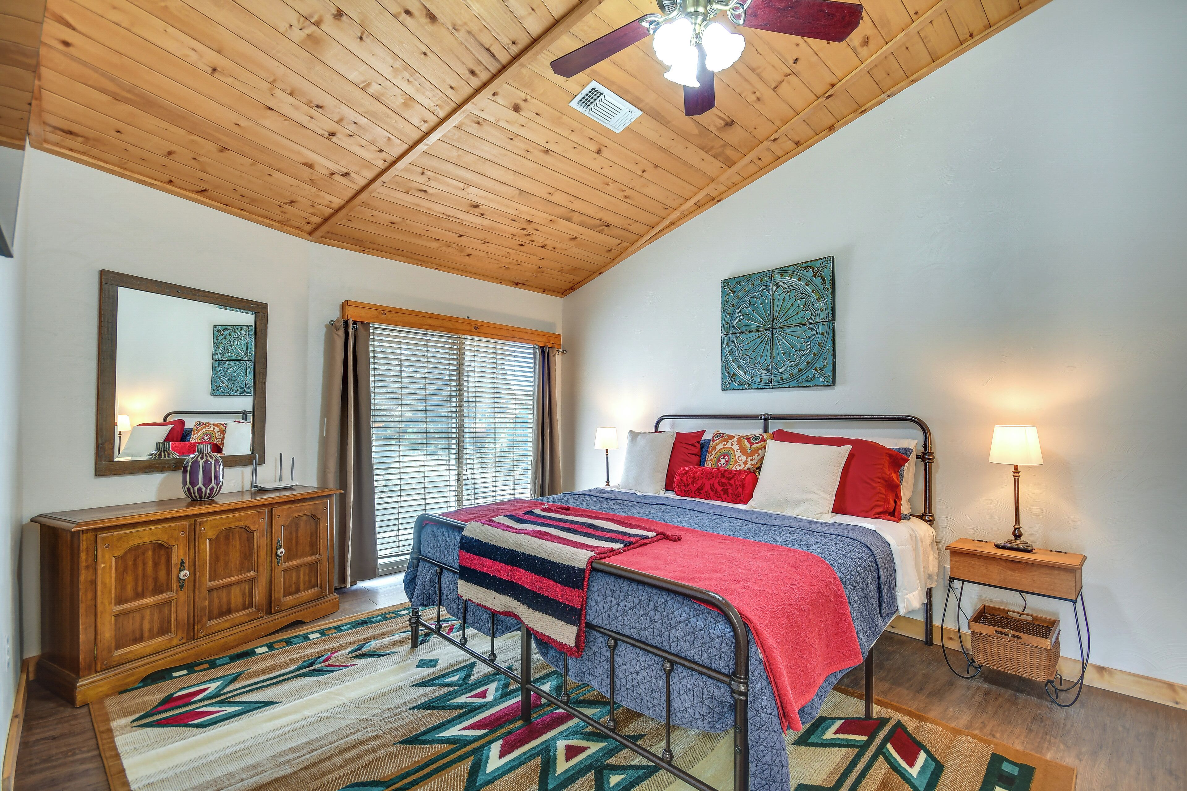 A bedroom with a wooden ceiling, a bed with a red and blue blanket, a nightstand with a lamp, and a large mirror on the wall.