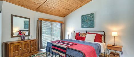 A bedroom with a wooden ceiling, a bed with a red and blue blanket, a nightstand with a lamp, and a large mirror on the wall.