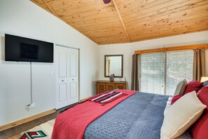 Quarto com teto de madeira, televisão na parede, cama com várias almofadas e cobertor, cômoda e janela com persianas.