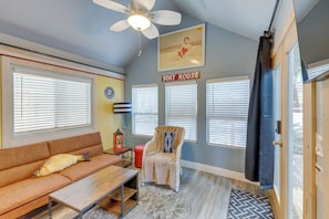 Una habitación con sofá, sillón, ventilador de techo y ventana con persianas.