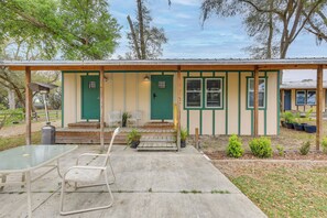 A small building with a green door and windows, a covered patio with a table and chairs, and potted plants.