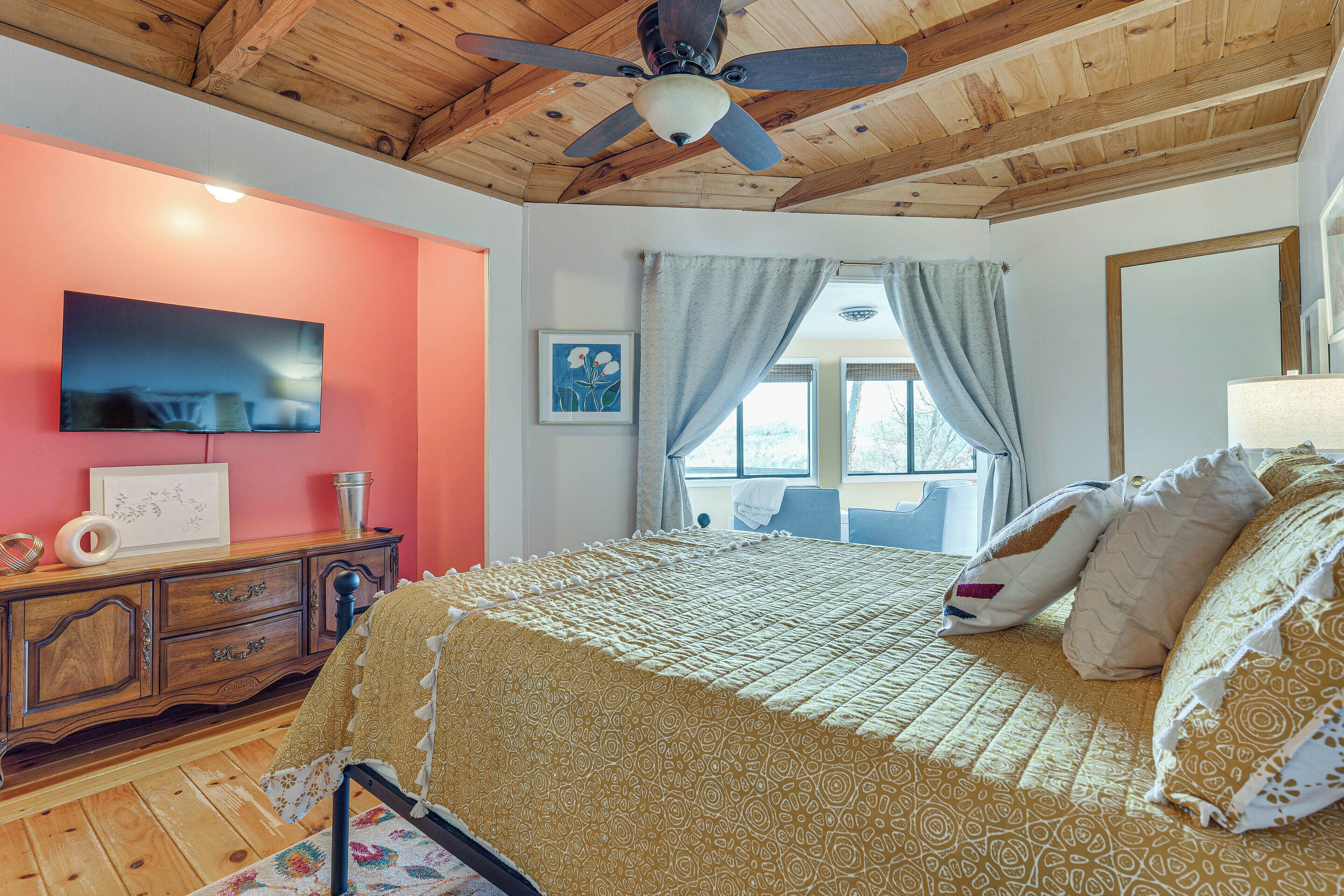 Une chambre avec un plafond en bois, un lit recouvert d’une literie à motifs, un téléviseur fixé au mur et une fenêtre avec des rideaux.
