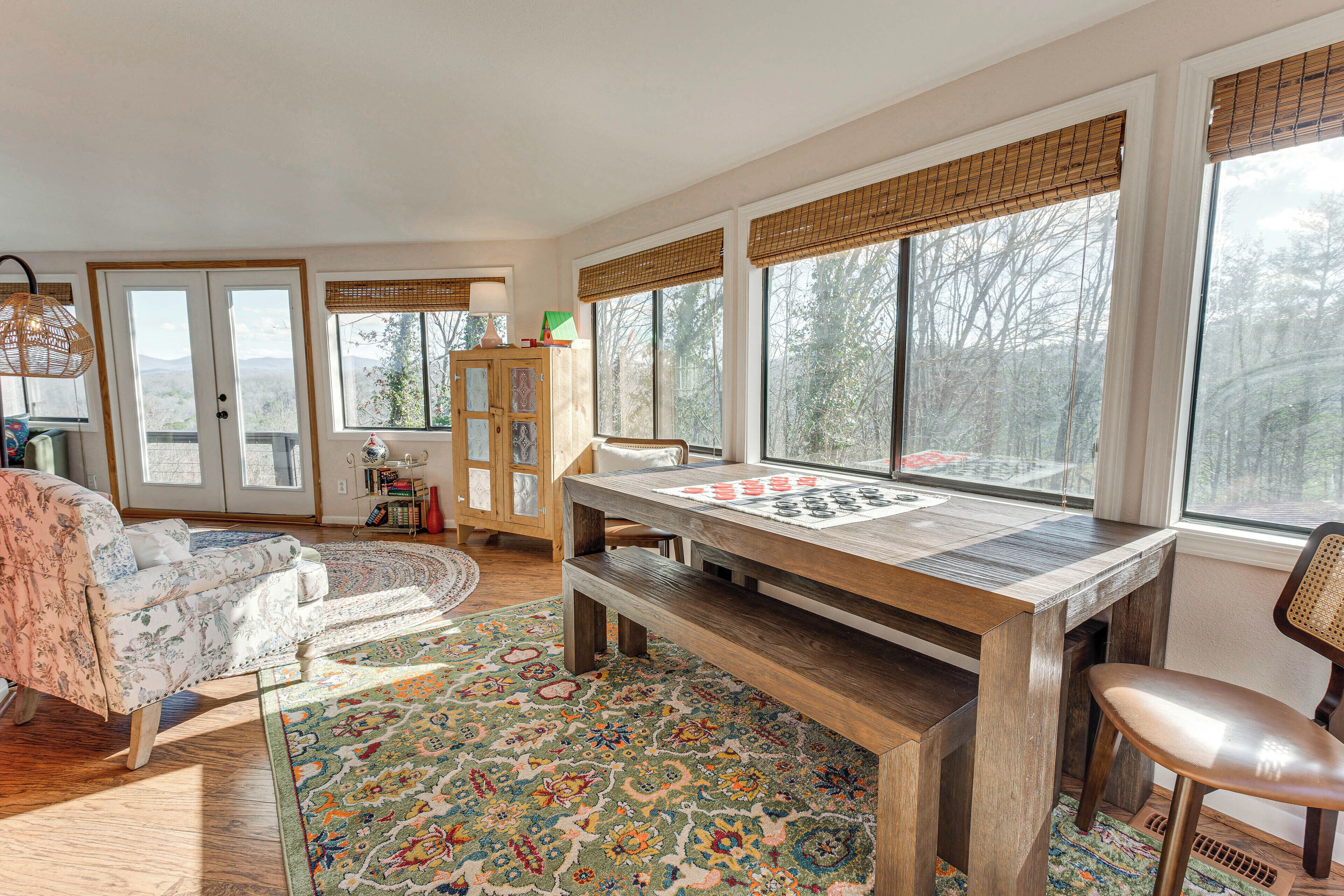 A spacious living room with a large wooden table, a sofa, and a chair. There are large windows with blinds, a bookshelf, and a rug.