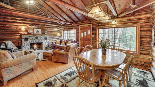 A log cabin interior with a stone fireplace, wooden furniture, and large windows offering a view of the outdoors.