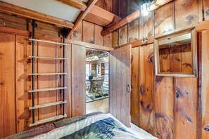 A wooden cabin interior with a bed, a wooden ladder, and a dining area visible through an open door.