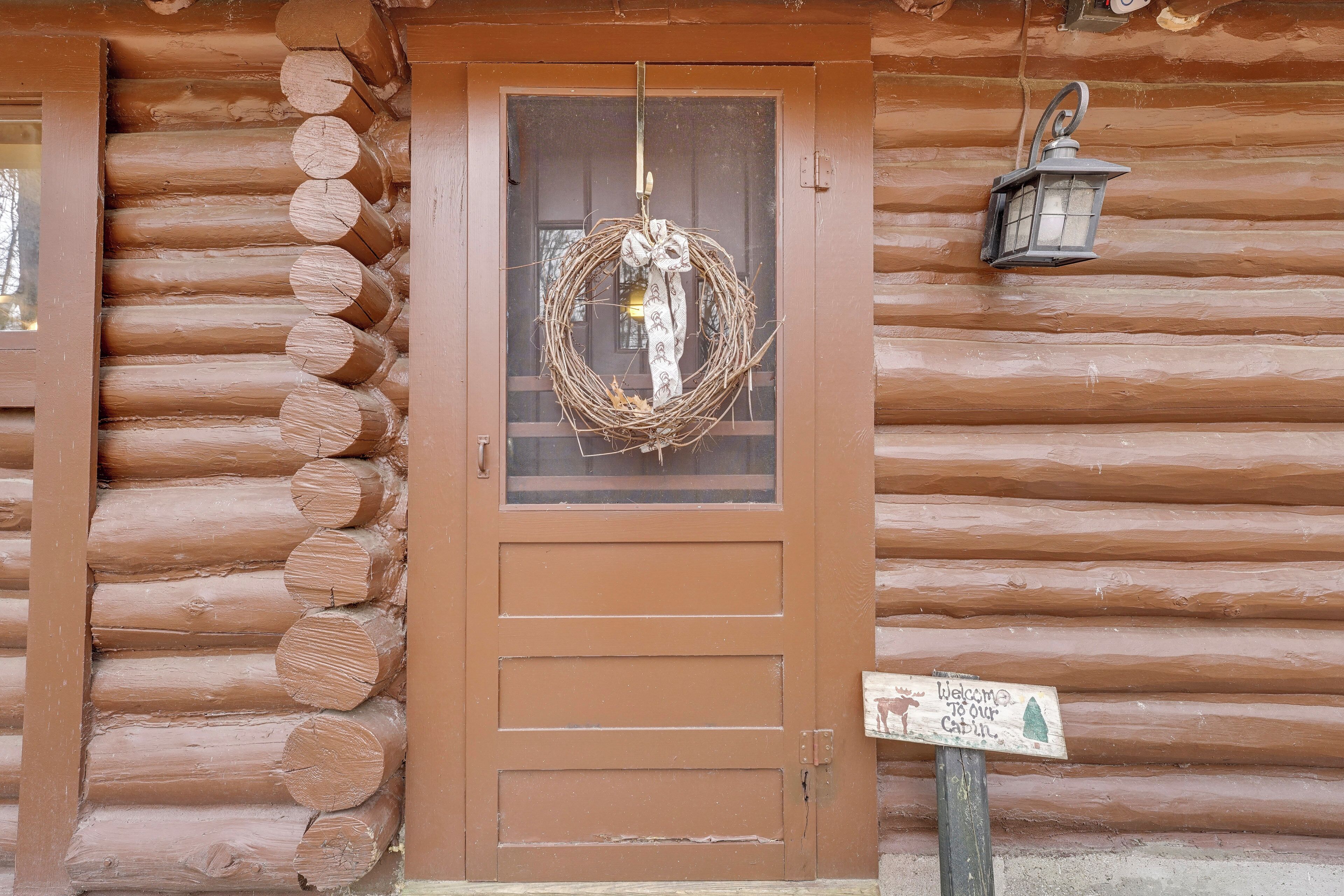 A log cabin with a brown door, a wreath hanging on the door, a lantern on the wall, and a welcome sign.