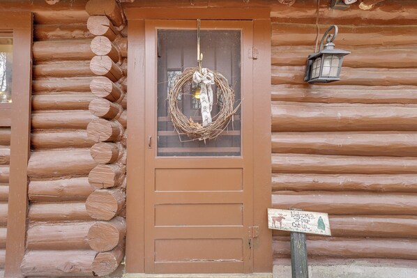 Una cabaña de madera con puerta marrón, un adorno colgando en la puerta, un farol en la pared y un cartel de bienvenida.