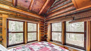A log cabin bedroom with a bed, large windows, and a view of the forest.