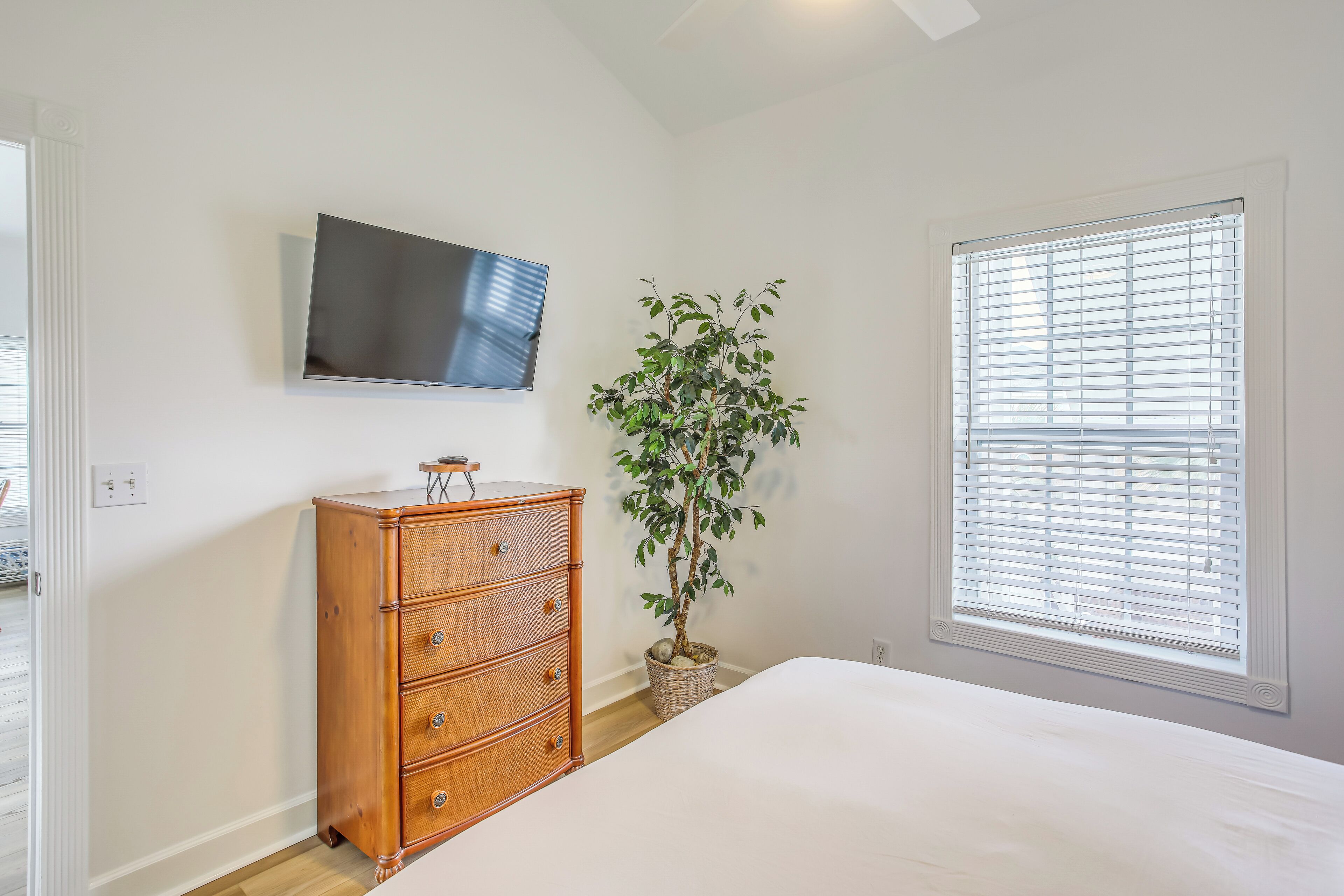 Un dormitorio con cama, cómoda, televisor, ventana con persianas y una planta en maceta.