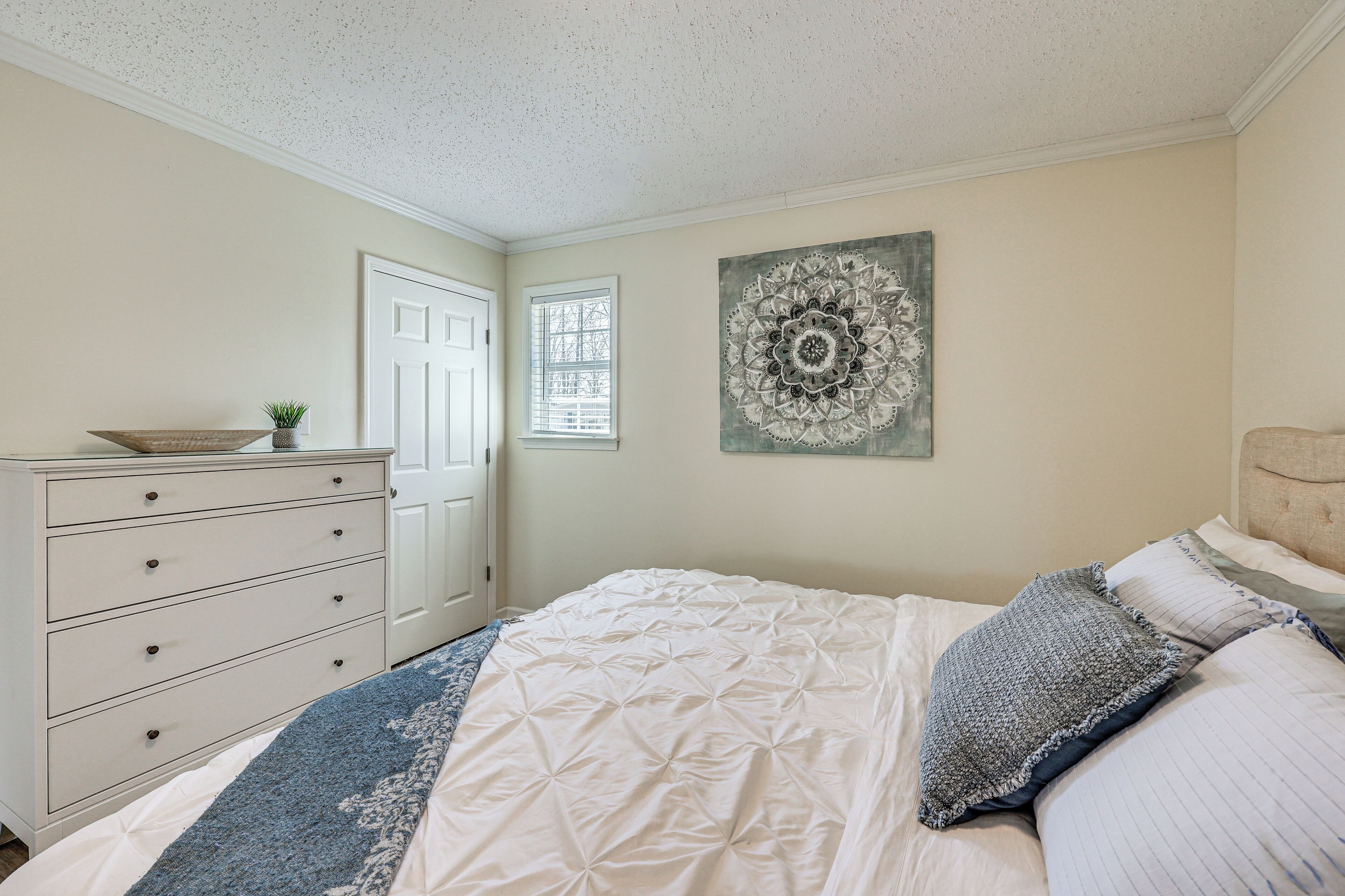 Um quarto com uma cama, penteadeira, janela e um enfeite na parede.