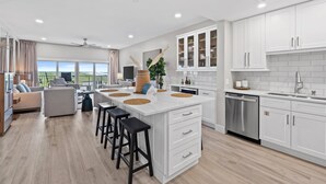 A modern kitchen with white cabinetry, a large island with stools, and a view of the outdoors. - Shell Seeker - Gulf-View Condo at Tops'l Resort | Pool, Beach Access, Hot Tub, & More! BM 702 (Miramar Beach)
