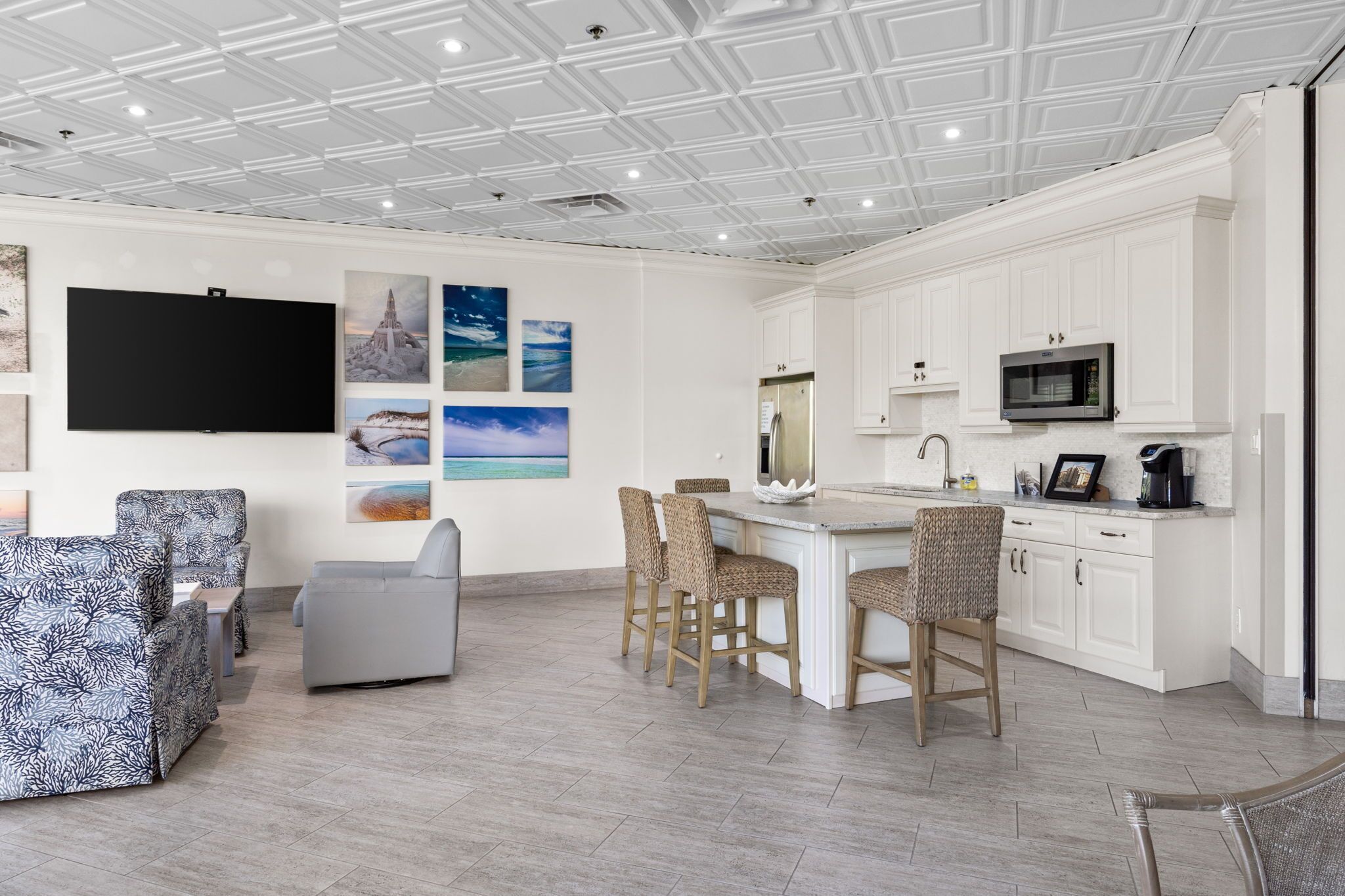 A spacious living area with a large TV, comfortable seating, a kitchen with white cabinets, and artwork on the walls.