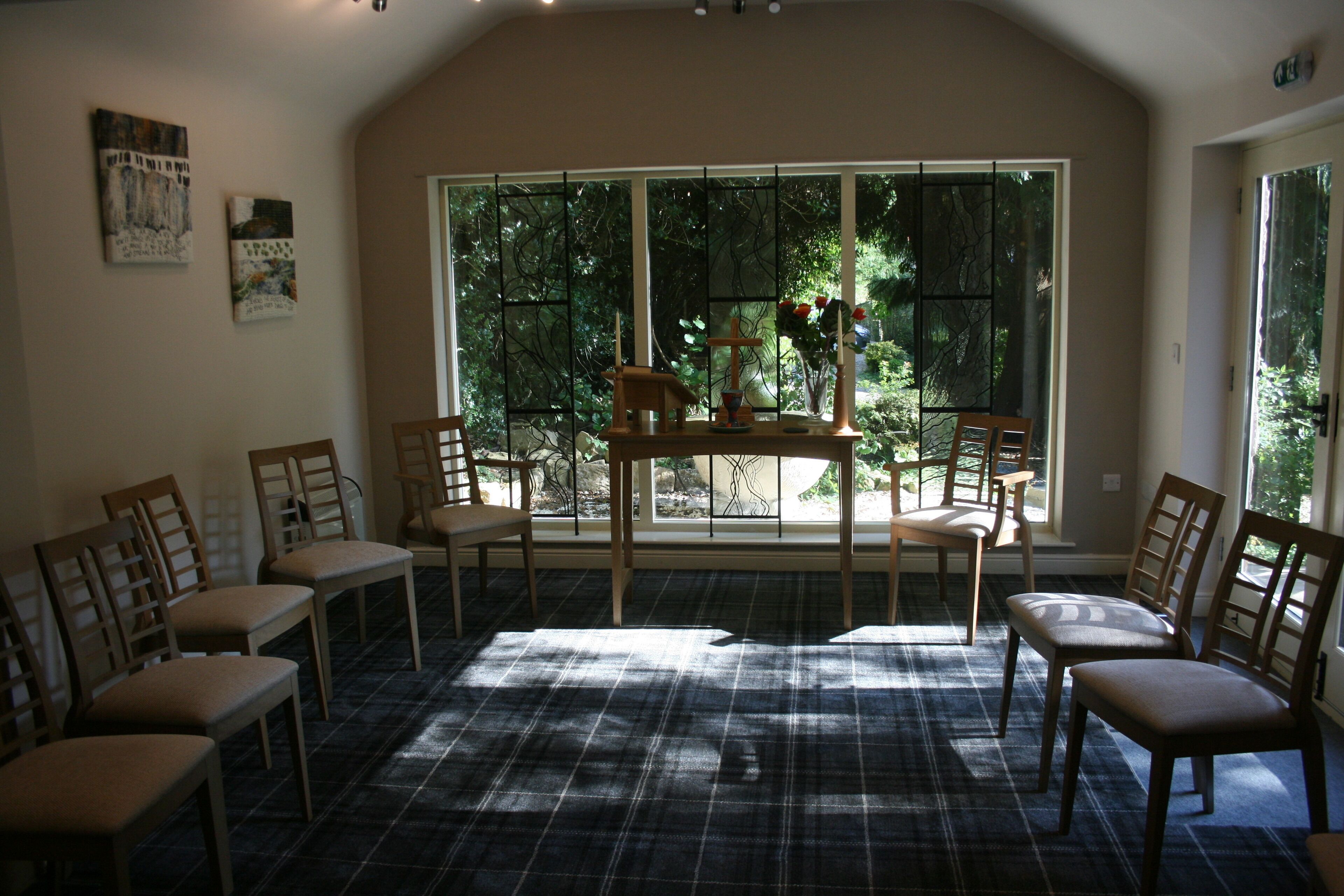 A room with a checkered floor, a table with a vase and a cross, and several chairs.