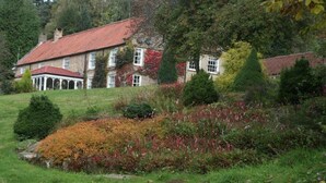 A large stone building with a red-tiled roof, surrounded by lush greenery and a variety of shrubs and trees. - High Dalby House (Pickering)
