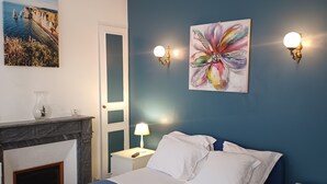 Un dormitorio con una cama azul, almohadas blancas, una mesita de noche con lámpara, una chimenea y dos luces fijadas a la pared.