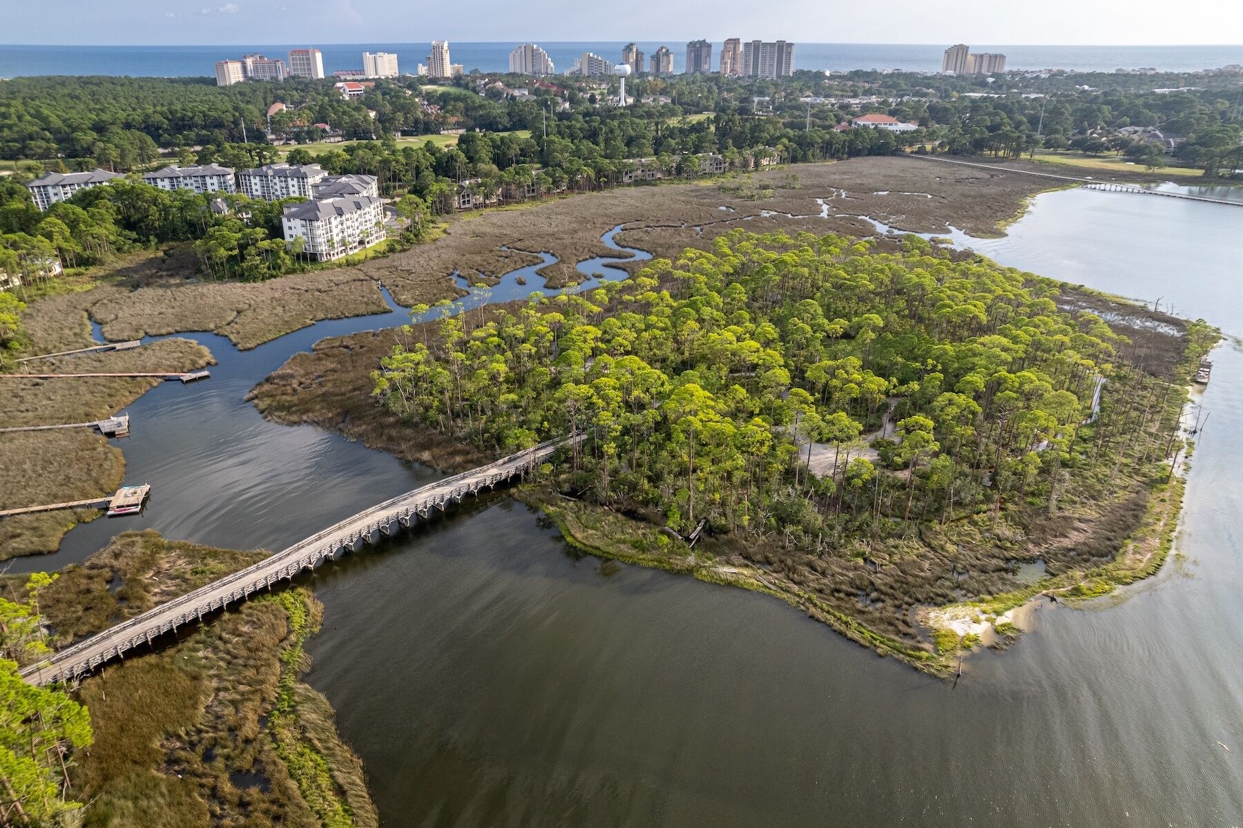 Une vue aérienne d’une zone côtière avec un pont reliant deux portions de terre, le tout entouré d’une végétation luxuriante et bordé par une étendue d’eau calme.