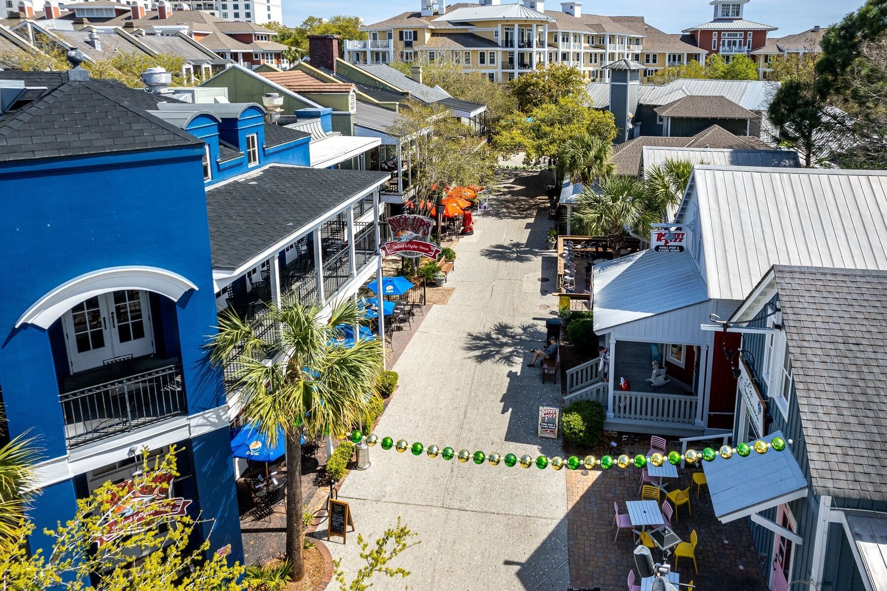 A street view of a coastal town with buildings, outdoor seating, and a row of shops.