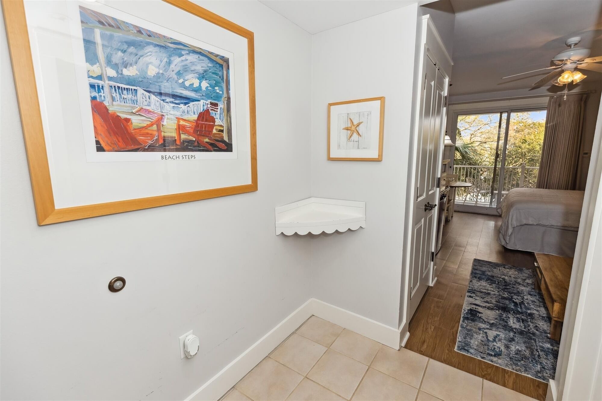 A hallway with a framed beach scene, a small white shelf, and a view of a balcony with a bed and a chair.