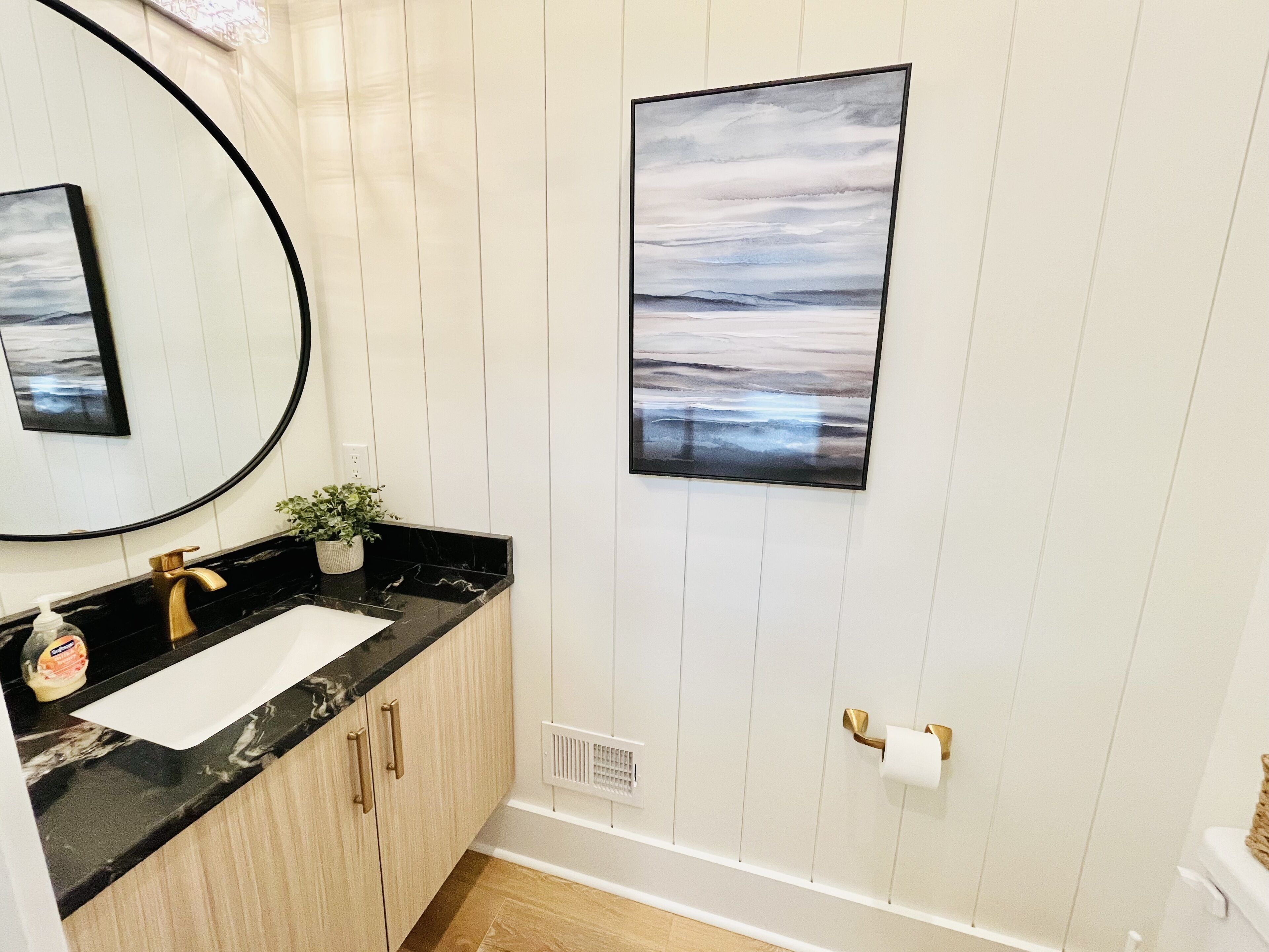 Een badkamer met een zwart aanrechtblad, een gouden kraan, een witte wastafel, een potplant, een lijst met een afbeelding en een toilet met een handdoekhouder.