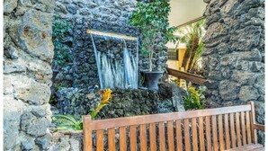 Eine Steinsäule mit Wasserfall, eine Holzbank und Kübelpflanzen.