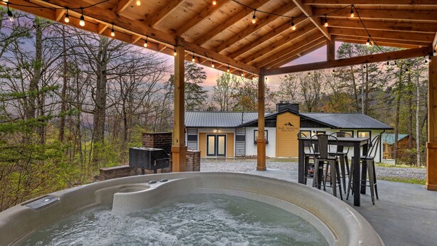 A hot tub under a wooden pergola with a view of a house and forest.