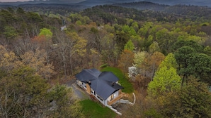 A house with a dark roof, surrounded by dense forest and mountains in the background.