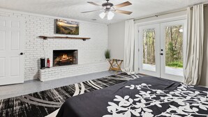 A bedroom with a white brick fireplace, a ceiling fan, and a large bed with a black and white patterned bedspread.