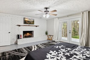 A bedroom with a white brick fireplace, a ceiling fan, and a large bed with a black and white patterned bedspread.