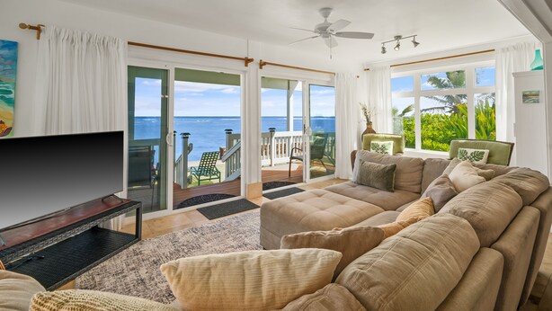 A spacious living room with a large flat-screen TV, comfortable seating, a ceiling fan, and a view of the ocean through large windows.
