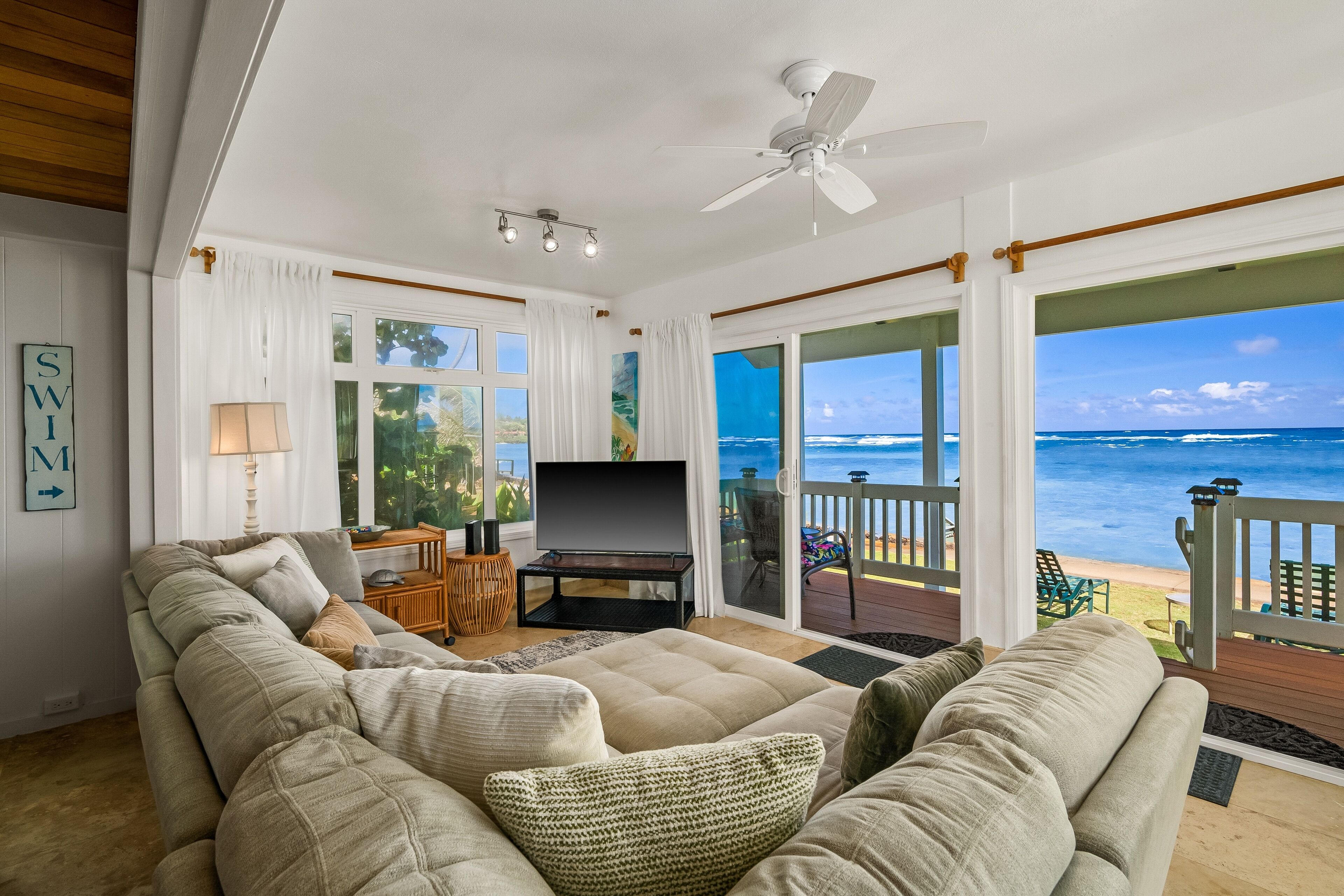 A living room with a large beige sectional sofa, a flat-screen TV, and a ceiling fan. There is a sliding glass door leading to a deck with a view of the ocean.