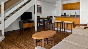 Una sala de estar moderna con una escalera de madera, un comedor con dos taburetes de bar y una cocina con armarios de madera y encimera.