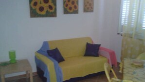 A yellow sofa with blue and black cushions, a small wooden table with a green object, and a yellow patterned chair. - Little house by the sea (Acquacalda)
