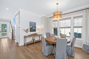 Een eetruimte met een ronde houten tafel en grijze gestoffeerde stoelen, een houten console met een lamp, en een schilderij van een pauw aan de muur.