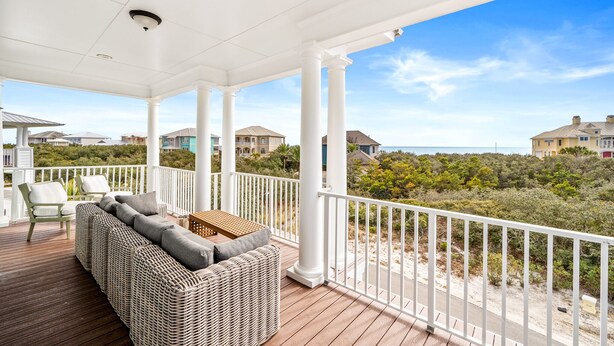 A covered outdoor area with a wicker sofa and chairs, white railings, and a view of houses and beach vegetation.