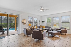 Un salon spacieux avec un grand canapé d’angle gris, plusieurs fauteuils, une table basse et un ventilateur de plafond. De grandes fenêtres laissent entrer beaucoup de lumière naturelle et offrent une vue sur l’extérieur.