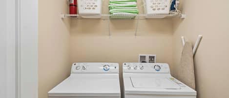 A laundry room with two white front-loading washers and dryers, a fire extinguisher, and a shelf with folded towels.