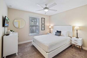 Une chambre à coucher comprenant un lit, des tables de chevet, un ventilateur de plafond, une fenêtre avec des stores, un téléviseur fixé au mur et des tableaux encadrés.