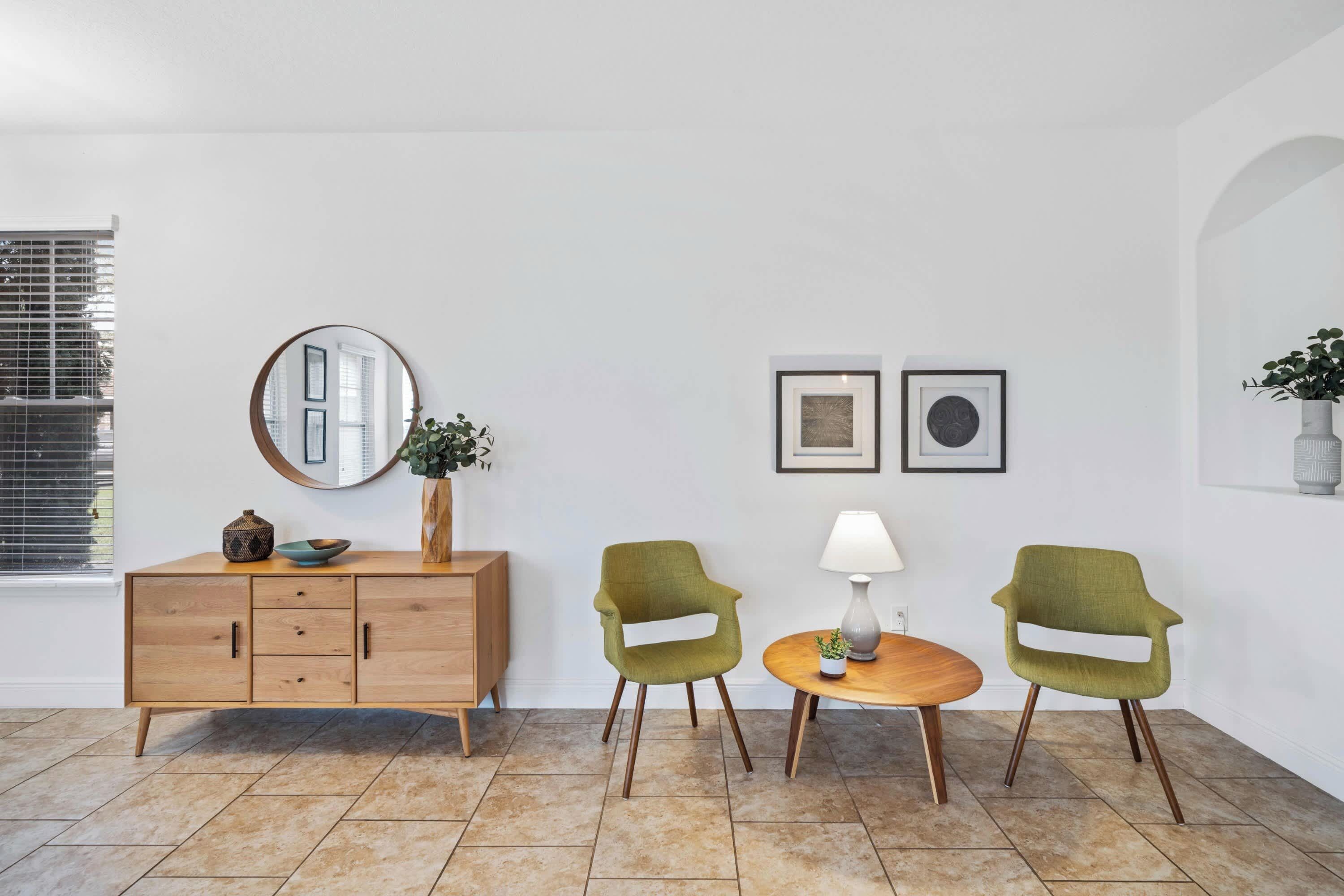 Une pièce comprenant un meuble en bois, deux chaises vertes, une table ronde, un lampadaire et deux tableaux encadrés accrochés au mur.