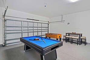 Une table de billard avec des billes et des queues, une table de baby-foot, et une petite table avec des chaises, le tout dans une pièce dont la porte de garage est blanche.