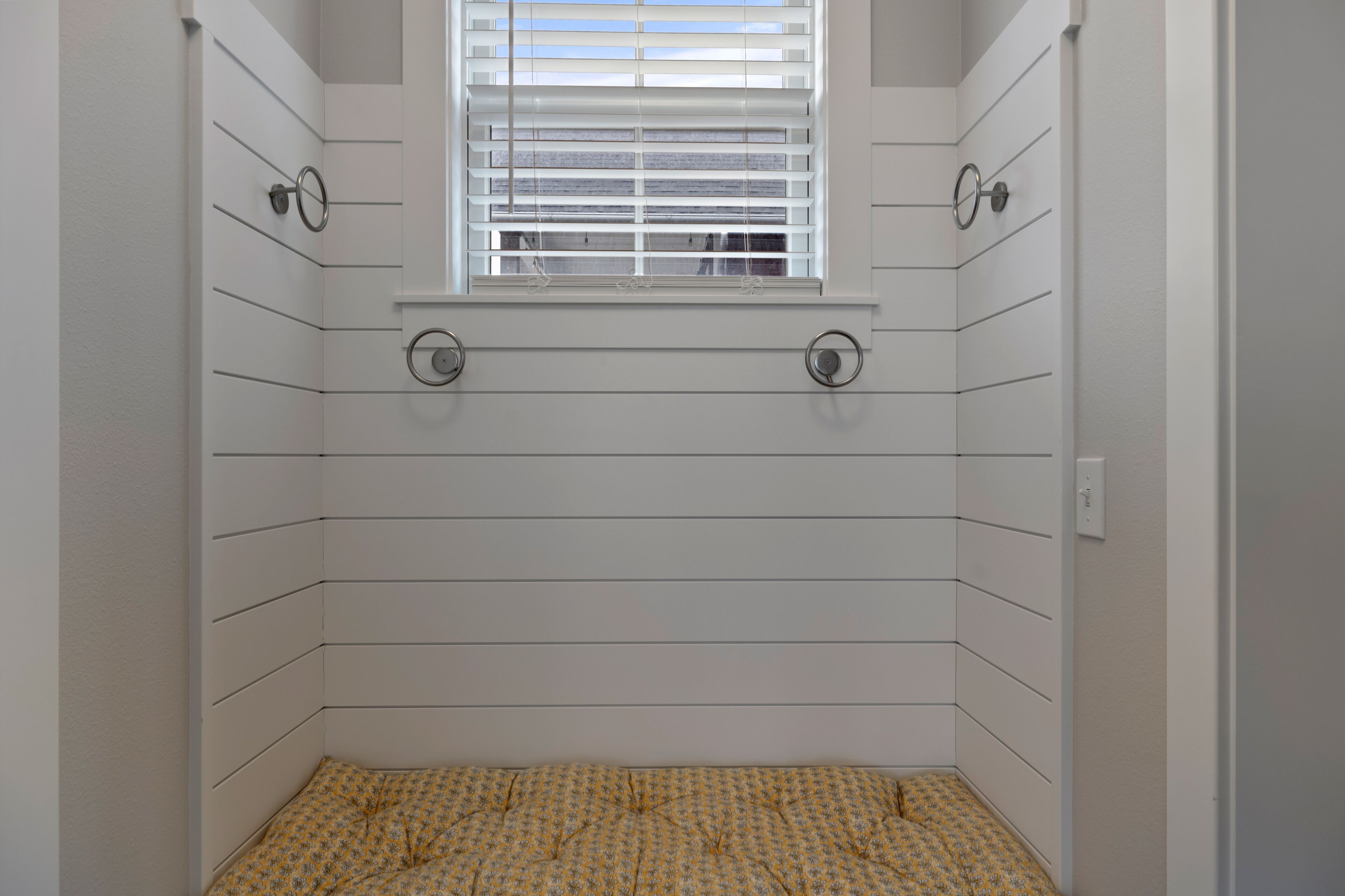 Une salle de bain avec des panneaux muraux blancs, une fenêtre avec des stores et un sol à motifs.